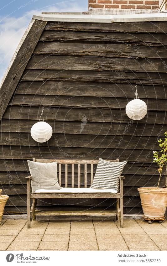 Wooden bench with pillows and lanterns hanging small cozy comfortable cushion white decor plant planter concrete floor tile wooden seat furniture empty wall