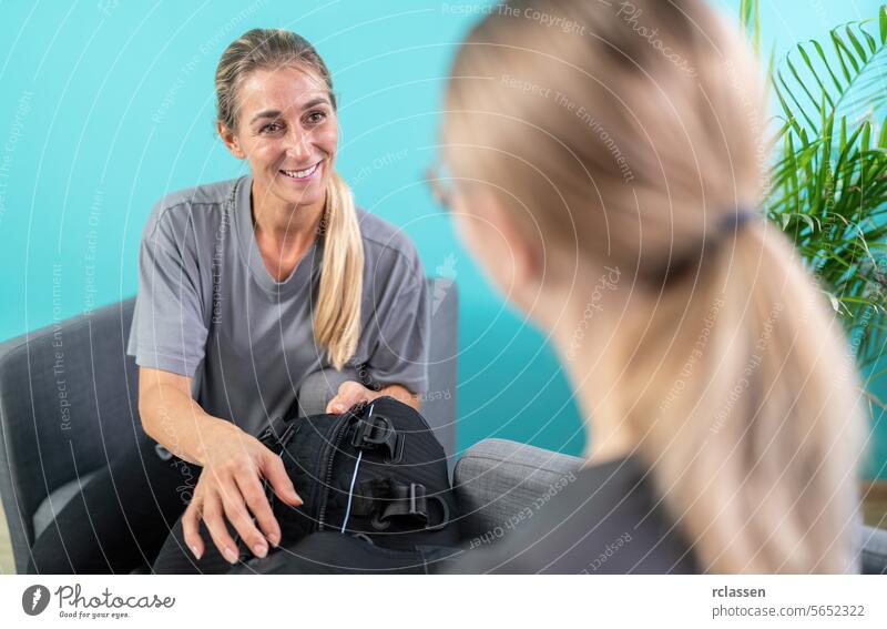 woman holding an EMS training suit while talking to a trainer in a consultation room ems training suit conversation armchairs indoor casual wear glasses fitness