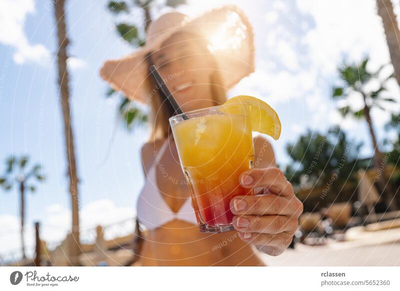 Close-up of a colorful cocktail held by a woman in a bikini, with a straw hat and palm trees at a caribbean island hotel party mallorca close-up colorful drink