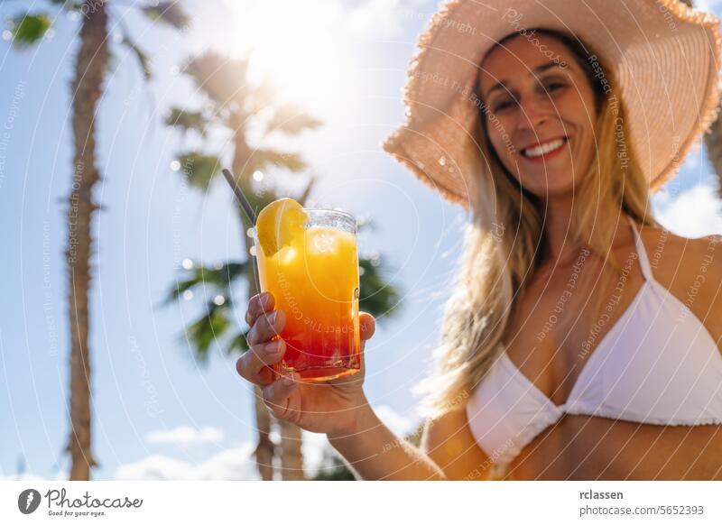 Woman in white bikini and straw hat offering a tropical cocktail, sunny sky with palm trees. Party and summer holidays concept image on a caribbean island.