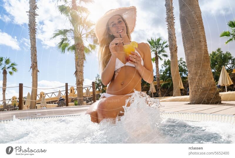 Woman in bikini with hat, drinking cocktail in whirlpool, palm trees and resort background at hotel in a island fuerteventura fun cheers woman sunny leisure