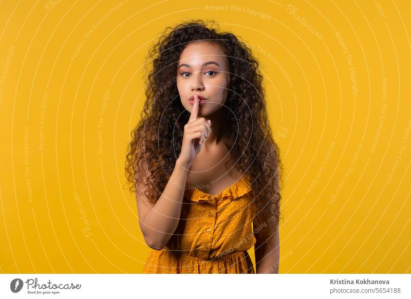 Smiling curly woman with finger on lips - shhh, secret, silence, yellow studio asking background biting charming closeup concept conspiracy contemplation cute