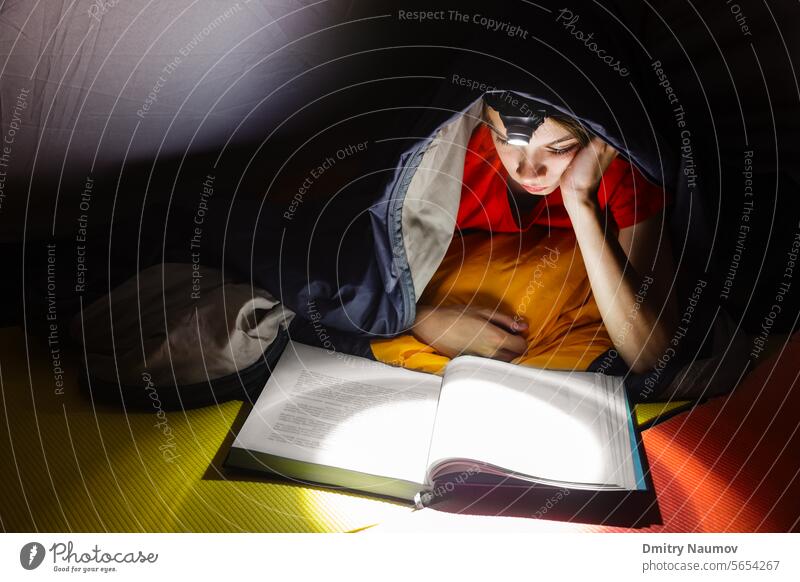 Teen boy laying in a camping tent wrapped in a sleeping bag reading a book with flashlight on at night Sleeping Bag bed bedtime calm child childhood