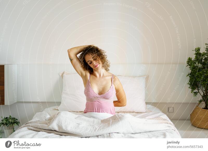 Happy woman doing stretching exercise on bed morning awake smile eyes closed nightwear cozy bedroom female adult happy glad comfort curly hair lady daylight sit