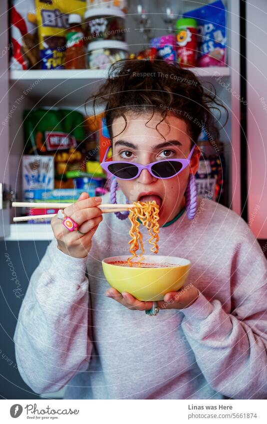 A stunning brunette, her curly brown hair framing her face, enjoys slurping spicy noodles in the comfort of her home. She exudes casual fashion while surrounded by a kitchen filled with candies. A model test of a teenage girl and some sweets. And noodles.