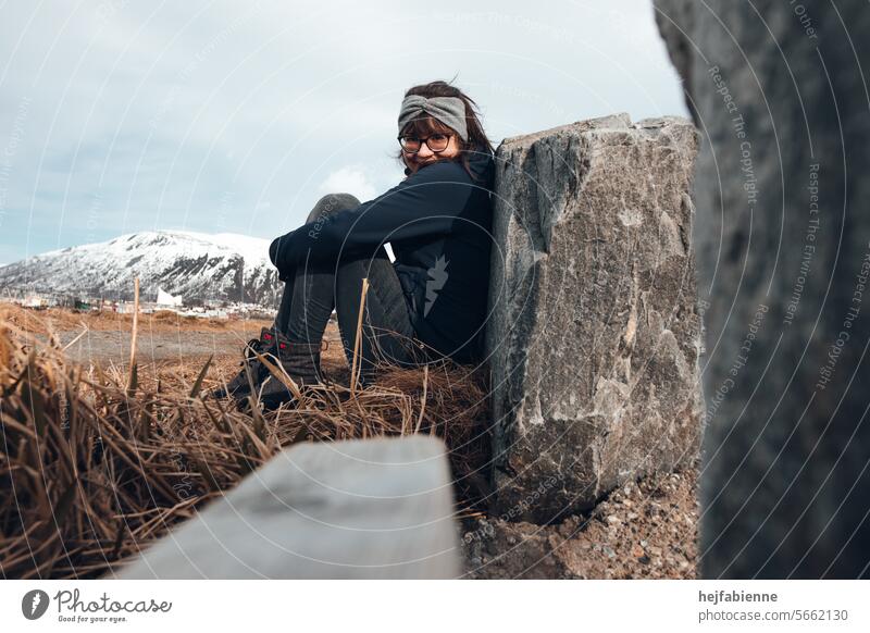 In the Arctic Circle: Portrait of a young woman in winter clothing and headband, sitting on the ground and leaning against a stone with her legs bent outdoor