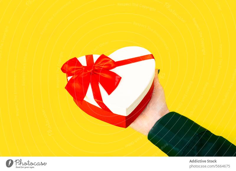 Anonymous hand holding a heart-shaped gift box with a red ribbon against a yellow background Hand valentine love celebration festive decoration elegant