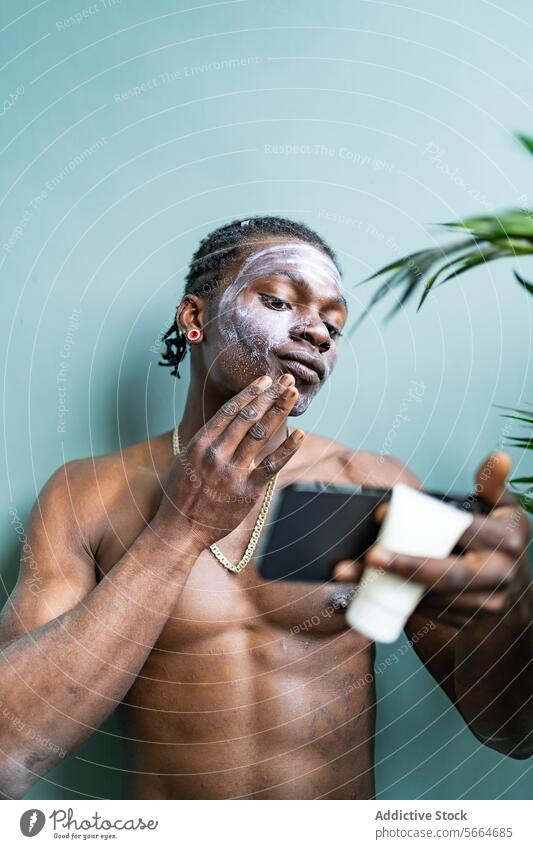 Black man applies facial mask while checking his reflection on a phone against a soft green background skincare black beauty health wellness adult young
