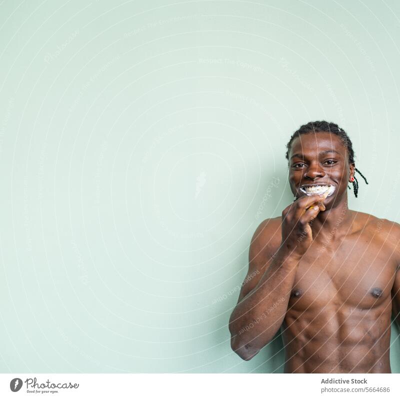 A shirtless black man with a nose ring and earrings brushes his teeth against a light green background dental care toothbrush hygiene toothpaste skin care