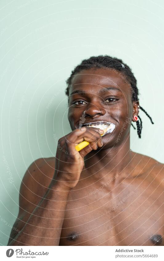 Portrait shirtless black man with a nose ring and earrings brushes his teeth against a light green background dental care toothbrush hygiene toothpaste