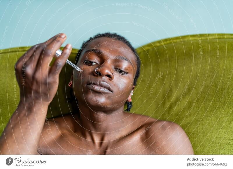 A black man applies serum to his face with a dropper while seated on a green sofa focusing intently on his skincare application facial care beauty health