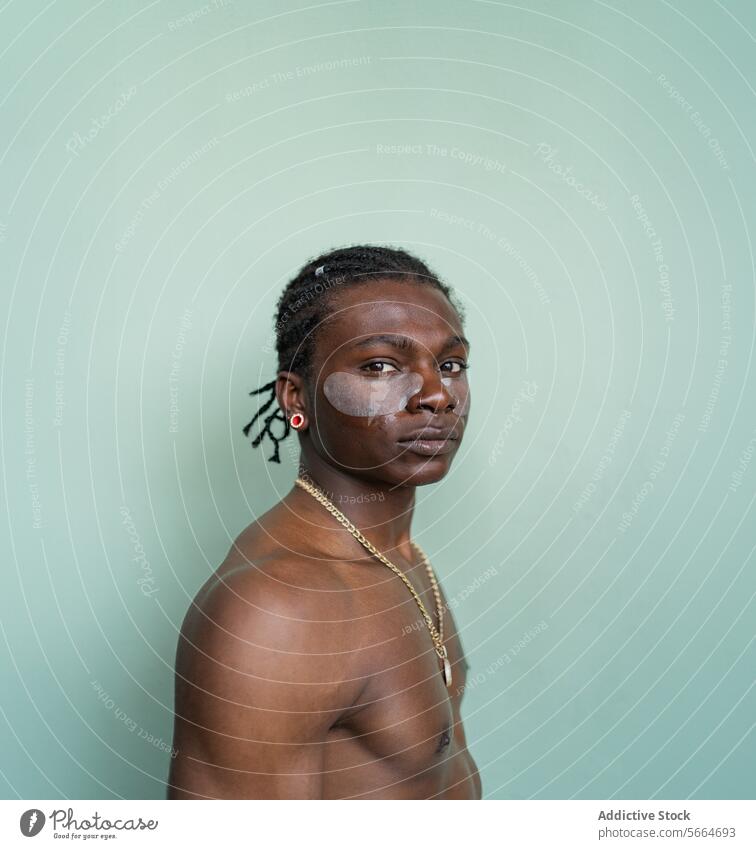 Side view of shirtless black man with facial cream on wearing earrings and a necklace stands against a light green backdrop skin care treatment green background