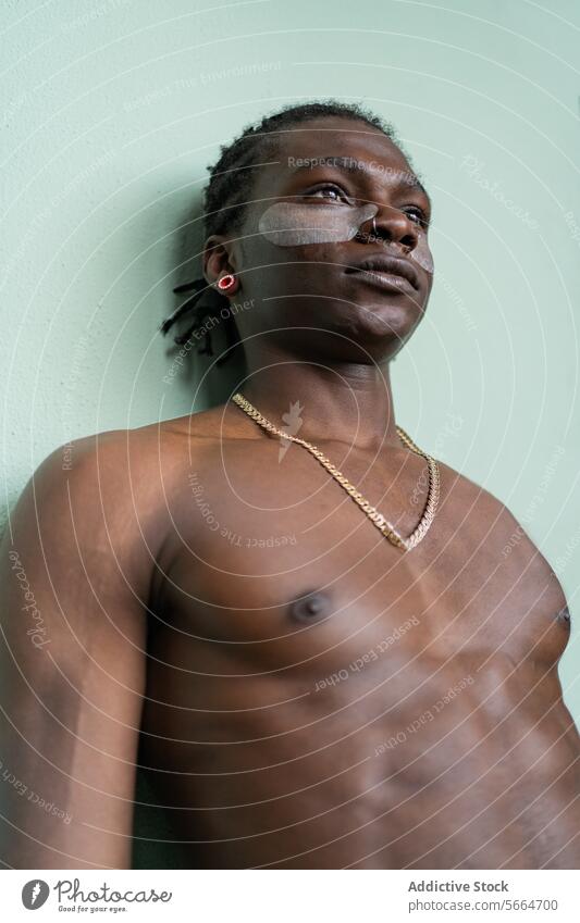 Low angle of a shirtless black man with under eye treatment patches wearing earrings and a necklace against a pastel green background skincare under-eye patch