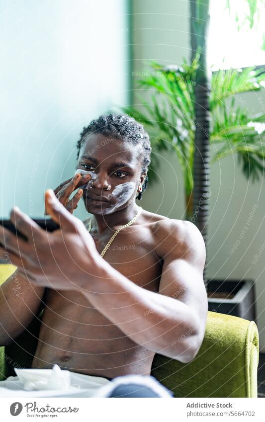 A focused black man applies a facial mask while looking at his reflection on a phone sitting indoors with plants in the background skincare application beauty