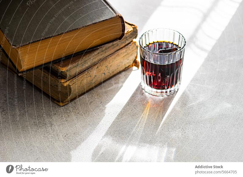 Cherry liqueur in a clear glass beside a stack of vintage books on a sunlit surface cherry sunlight shadow alcohol drink beverage bright table reflection red