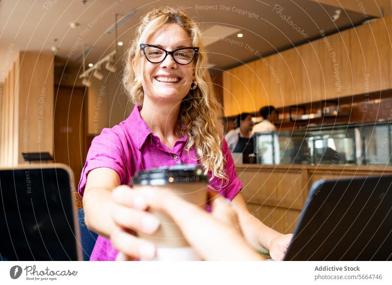 Caucasian cheerful blonde woman with glasses handing a coffee cup, sharing a moment with a colleague in a cafe during remote work environment in Chiang Mai, Thailand