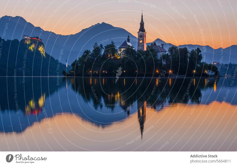 The sun sets on Lake Bled, casting a warm glow on the Pilgrimage Church of the Assumption of Maria and the Bled Castle Sunset Slovenia dusk twilight reflection