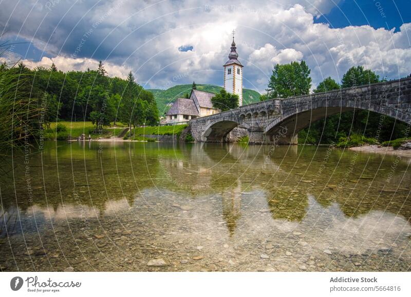 A stone bridge arches over the crystal-clear waters of Lake Bohinj with the Church of St. John the Baptist and mountains in the backdrop Stone church Slovenia