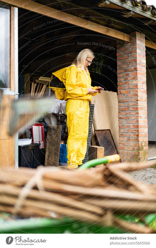 Focused female Beekeeper in yellow overall using mobile phone while working in apiary against building woman smartphone beekeeper focus gadget mature apiculture