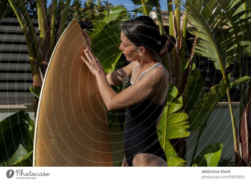 Side view of focused female surfer touching a surfboard while standing by tropical foliage in the garden Woman Female preparation outdoor backyard summer