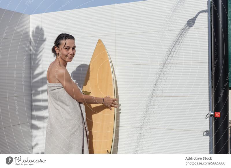 Smiling woman in a towel standing looking at camera next to a surfboard after a shower Woman smiling outdoor summer leisure relaxation backyard swimsuit