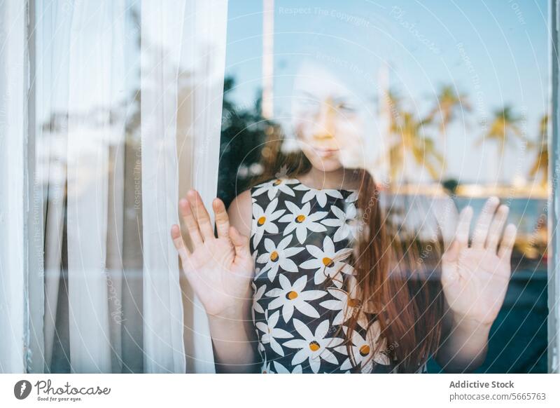 Young woman looking through a window with palm trees reflection thoughtful gaze glass hand press blur background tranquil environment young female contemplation