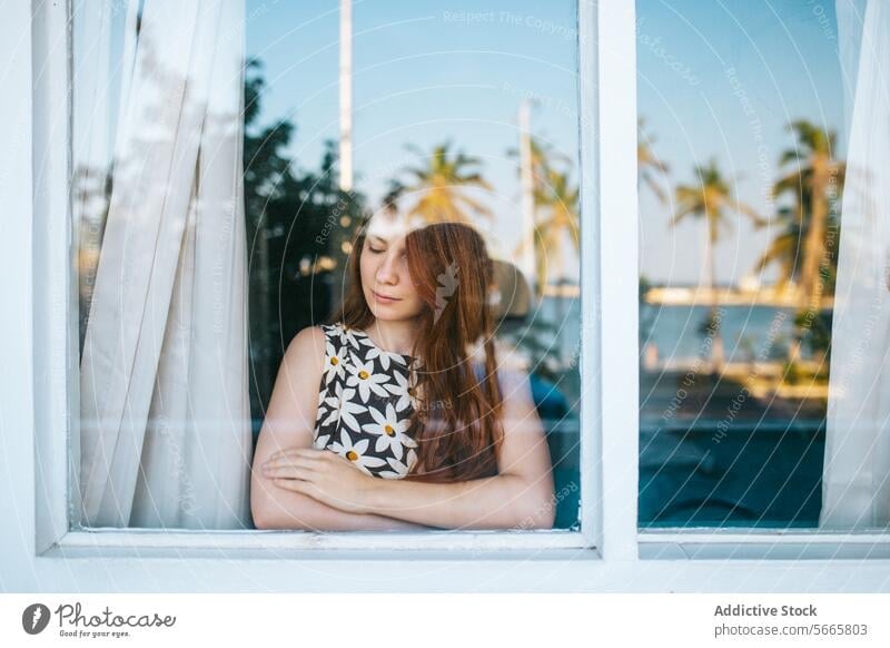 Contemplative woman looking out a window contemplation thoughtfulness reflection gazing serene palms background young arms crossed indoors blurred background
