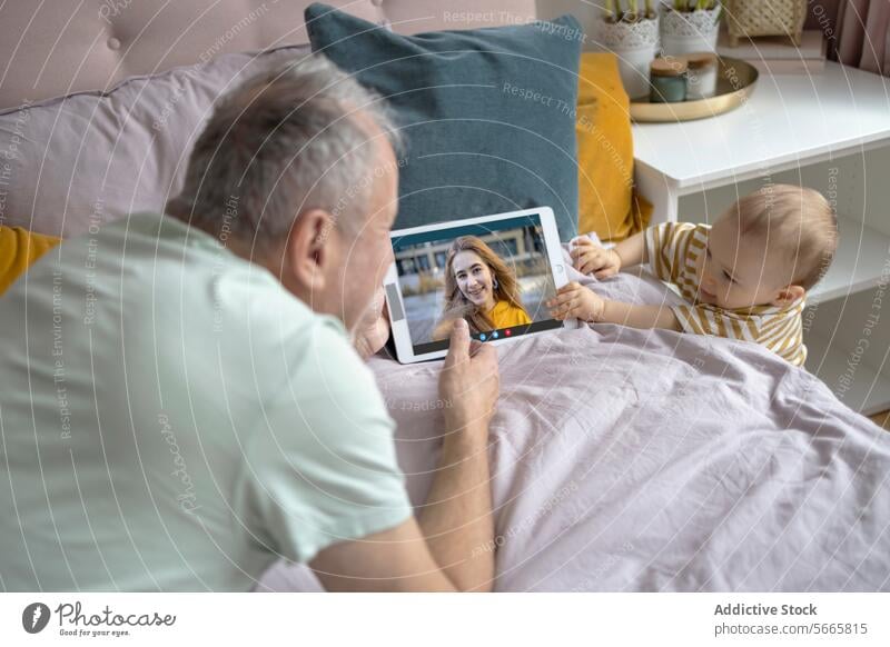 Grandfather and baby video calling with family on tablet grandfather bed pillows comfort digital technology infant senior man young woman sitting cozy bedroom