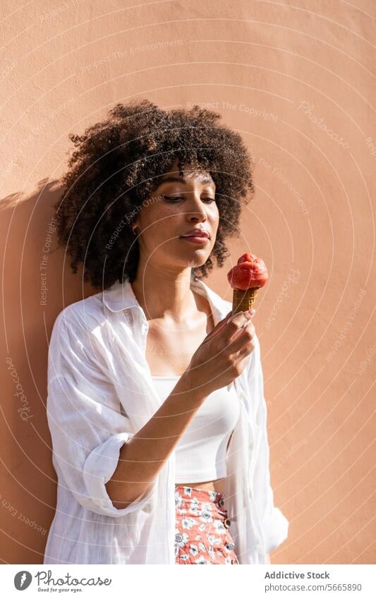A woman with curly hair savors a strawberry ice cream cone against a peach-colored wall embodying a relaxed summer vibe Woman peach wall portrait enjoying
