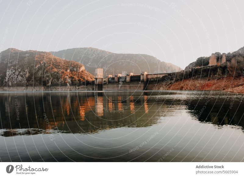 Tranquil waters reflect a dam surrounded by rugged mountains during a calm sunset highlighting the harmony of industrial and natural landscapes reflection