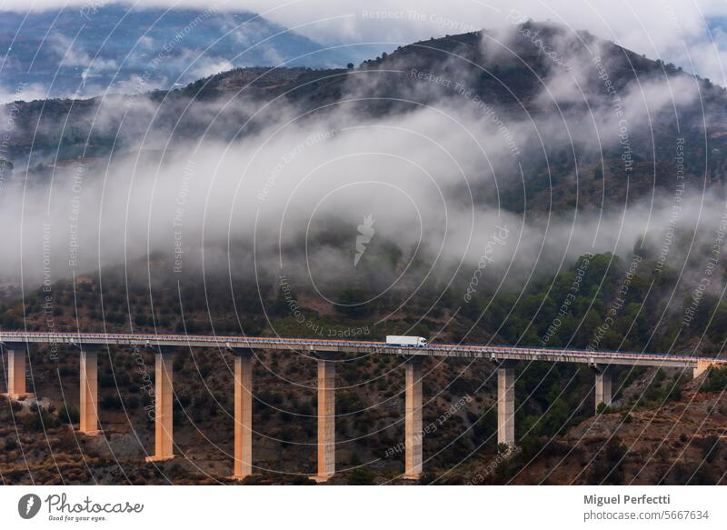 Truck with a refrigerated semi-trailer driving over a bridge and a mountain landscape with low clouds in the background. truck transport viaduct highway sunset