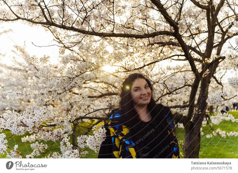 Young attractive plus size woman with long dark hair gets pleasure spending time in blooming cherry orchard in park young background trees city spring urban