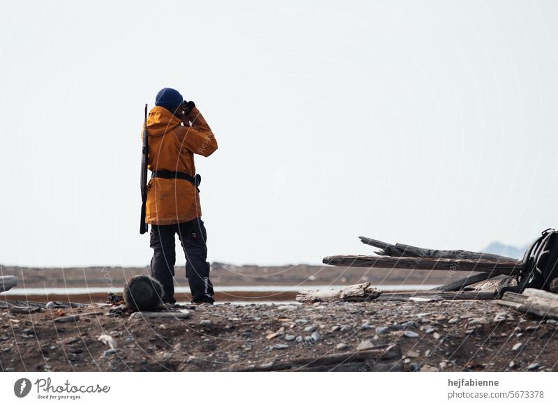 Polar bear guard on Spitsbergen during an expedition. Female guide with yellow ranger jacket and rifle in the Arctic tundra monitors the surroundings with binoculars.