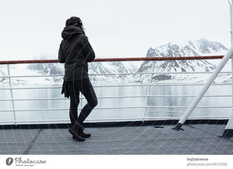 A young woman in winter clothing stands at the railing of a cruise ship and looks out over an Arctic fjord during a Spitsbergen expedition. Young woman