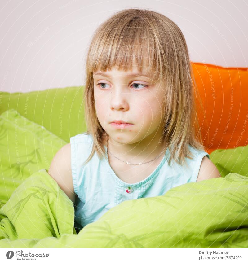 Portrait of upset 6 year old blond girl sitting in her bed with a chickenpox rash on her face afflicted blister bored chicken pox child childhood condition