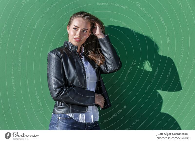 Confident young woman adjusting hair against green wall shadow sunlight fix long hair appearance leather jacket touch hair confident smile positive self assured