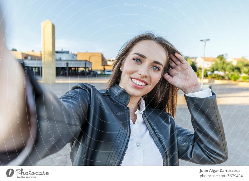 Blue-eyed blonde woman in leather jacket smiling takes a selfie with her camera in the street portrait touching her hair happiness hairstyle front view