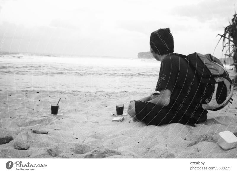 Young adult man sitting alone on sand and looking far away on side at sea beach with a glass cup of coffee and daypack bag on the back person young vacation