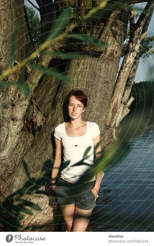 Portrait of a young, slim, beautiful woman with freckles in a white top and shorts, leaning against a tree and slightly covered by a branch with leaves Woman