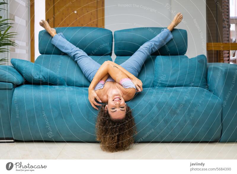 Cheerful woman lying upside down on sofa at home having fun happy living room smile relax laugh curly hair couch enjoy optimist glad content positive lounge