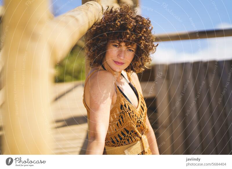 Young curly haired lady standing on coast woman lean on path shore idyllic pleasure season sunlight resort beach smile positive sensual female young brunette