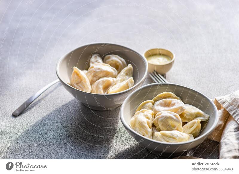 A pair of ceramic bowls containing Ukrainian varenyky, traditional dumplings, served with a side of sour cream and butter sauce, illuminated by natural light