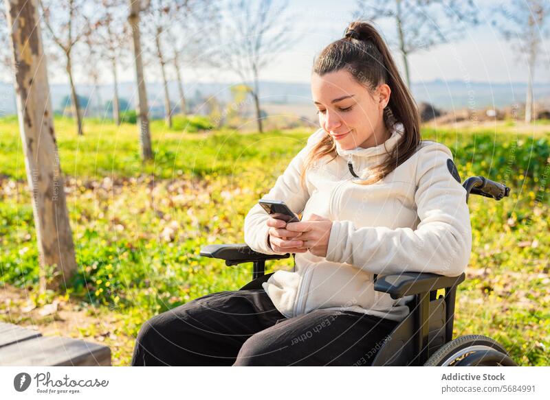 A young woman in a wheelchair is smiling while browsing on her smartphone in a park Woman bench mobile phone outdoor casual wear sitting female technology