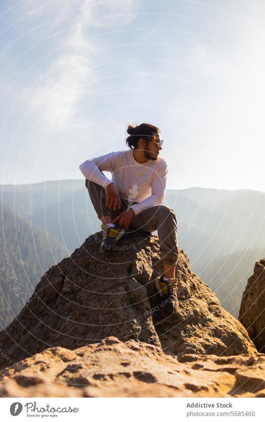 Young man sitting on cliff near foggy mountains admire tourist casual outfit beard sunglasses rocky nature peak traveler trip explore journey adventure male