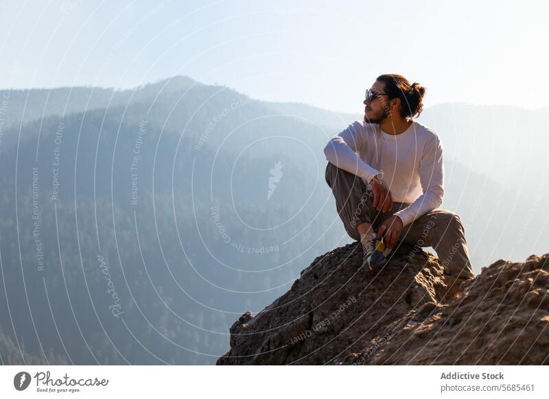 Young man sitting on cliff near foggy mountains admire tourist casual outfit beard sunglasses rocky nature peak traveler trip explore journey adventure male