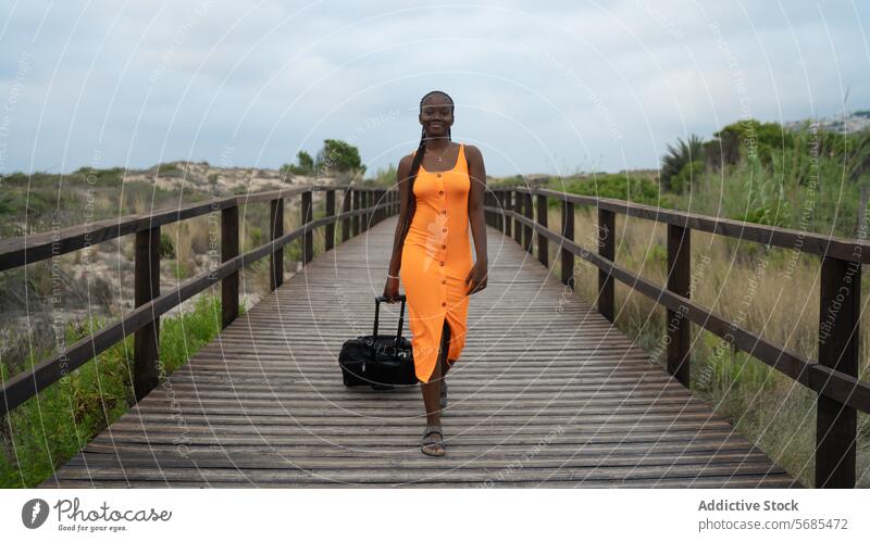 Smiling black woman walking on wooden footbridge lady gown drag luggage smile positive traveler hill mountain nature vacation female bush boardwalk trip dress