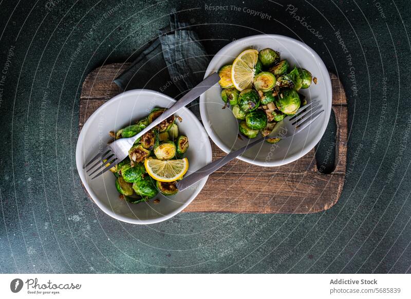 Top view of bowl of grilled Brussels sprouts seasoned with garlic and spices, garnished with a slice of lemon, served on a wooden board with a fork Bowl food