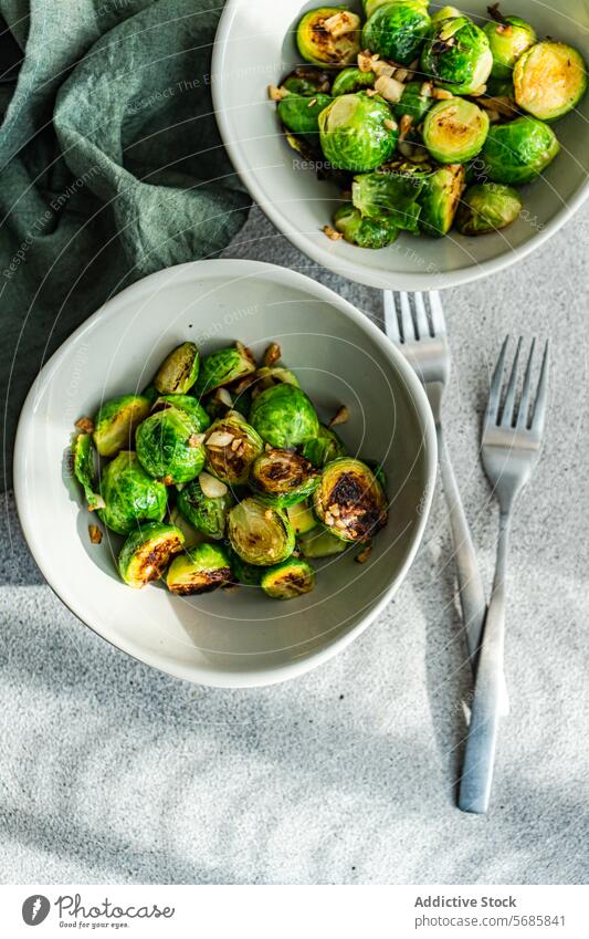 Top view of two bowls filled with barbecued Brussels sprouts flavored with garlic and spices, accompanied by silver forks on a textured tablecloth Bowl healthy
