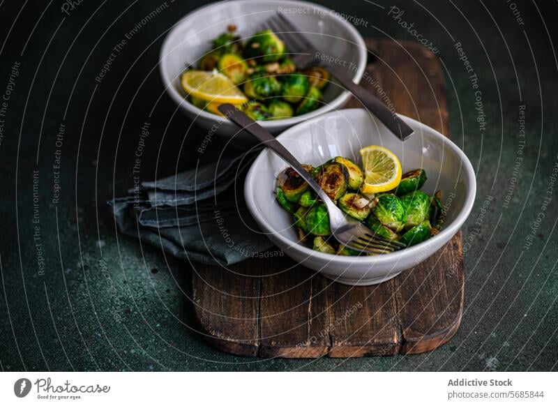 From above of bowl of grilled Brussels sprouts seasoned with garlic and spices, garnished with a slice of lemon, served on a wooden board with a fork Bowl food