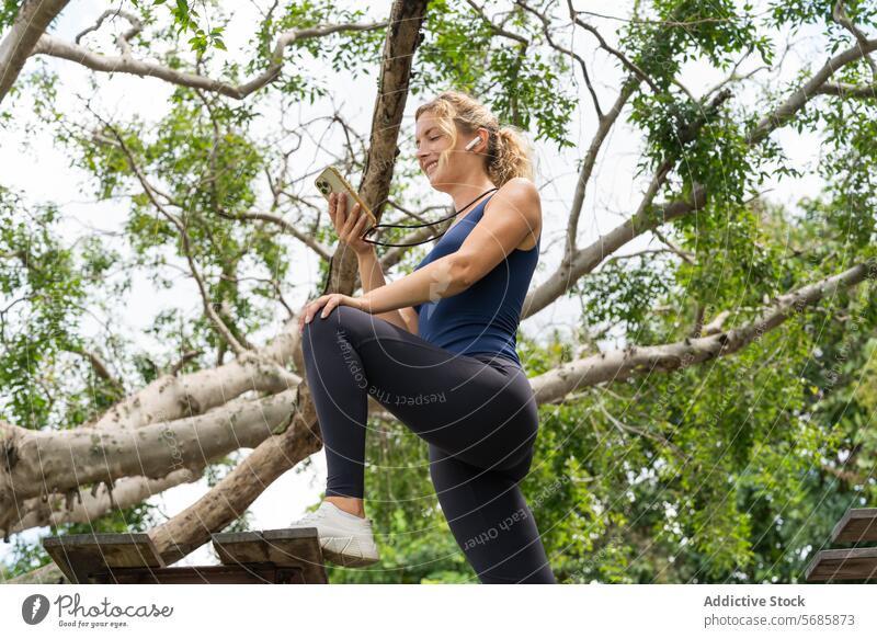 Smiling woman using smartphone near tree branches fitness training plank slim activewear sneakers mobile smile workout sportswoman exercise gadget wellbeing